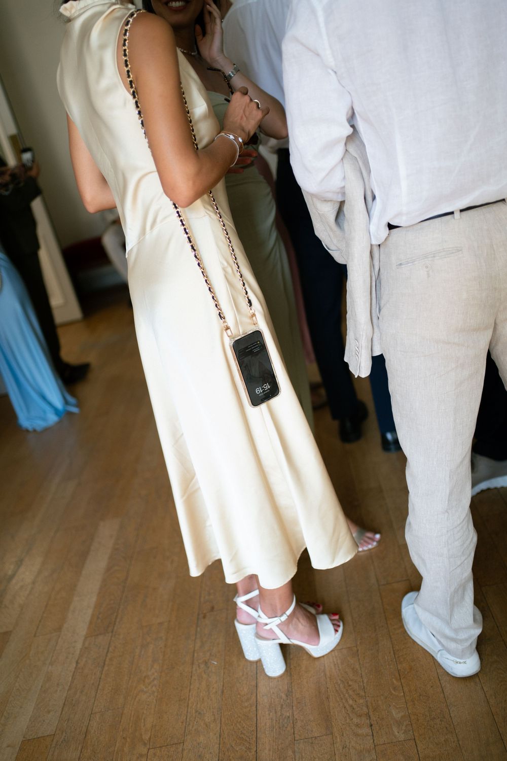 Bride in ivory midi dress with crossbody bag and white heels stands beside groom in linen suit.