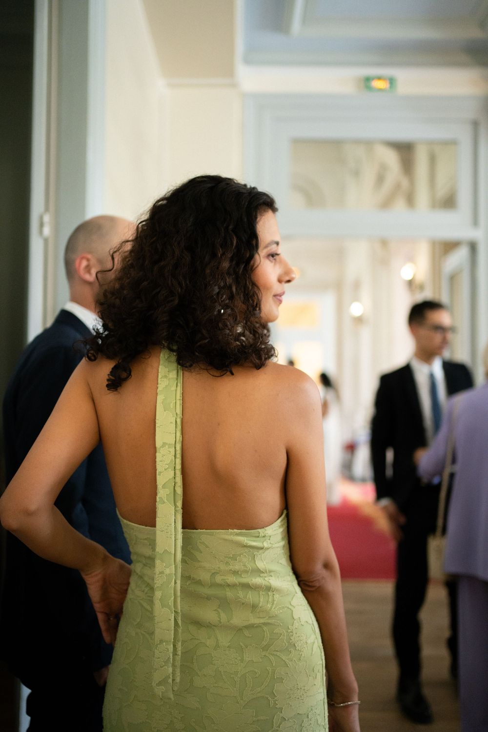 Woman in backless green halter dress with curly hair stands at elegant indoor event venue.