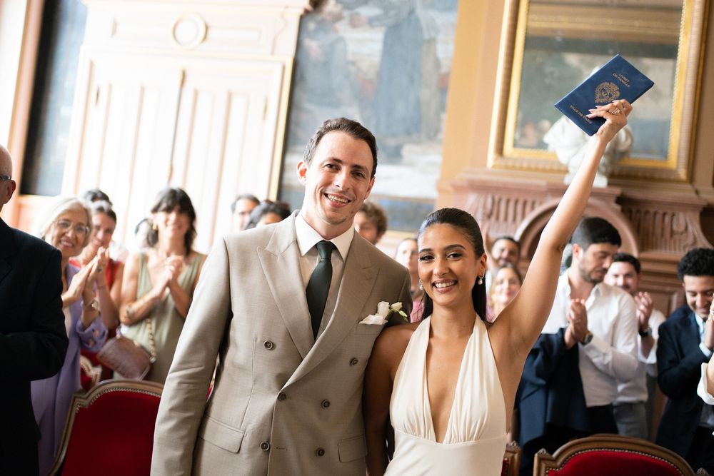 Joyful newlyweds celebrate after wedding ceremony, bride raising marriage certificate, guests applauding in elegant venue.