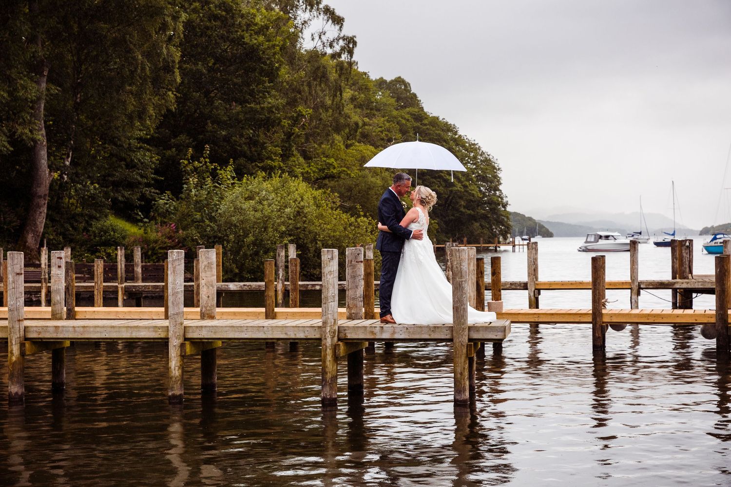 Bride and groom share umbrella on rainy lakeside dock with boathouse and sailboats in background.