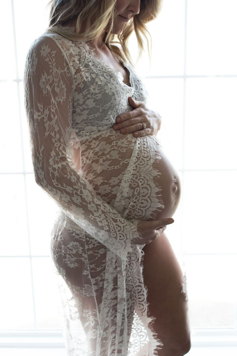 Artistic maternity photo showing silhouette in white lace dress against window light.