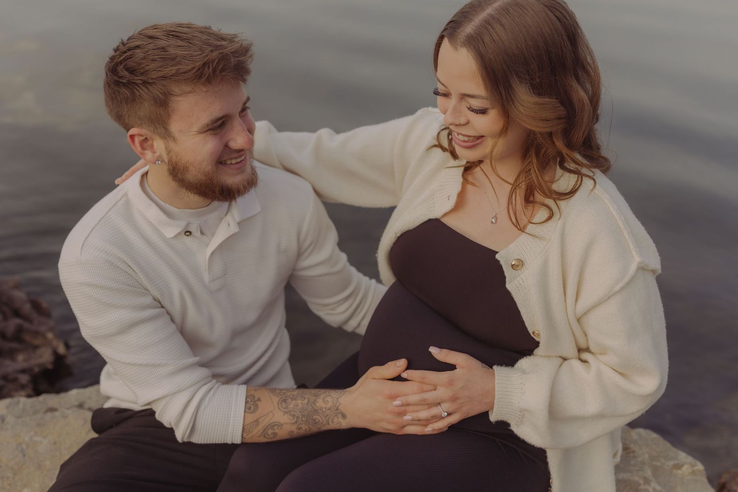 Couple sitting on rocks by water, man smiling at pregnant woman in cream cardigan and black outfit.