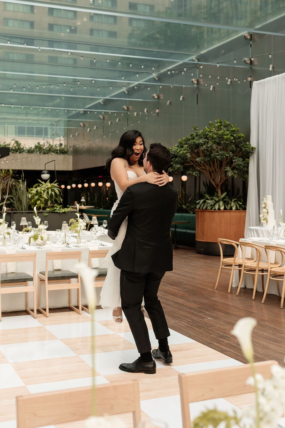 Bride and groom share a joyful first dance on a checkered floor at an elegant indoor wedding reception. Captured by Aliza Dworkind.