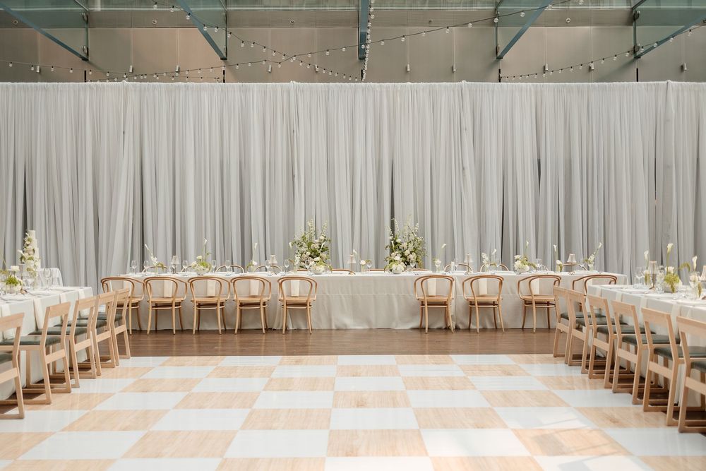 Elegant wedding reception with white drape backdrop, long tables, wooden chairs, floral centerpieces, and checkered dance floor. Le Cathcart