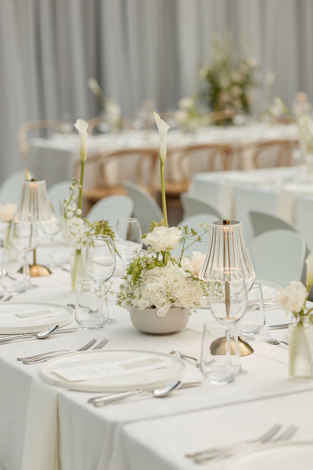 Elegant wedding reception table with white floral centerpieces, gold candle holders, and fine dining settings on white linen.