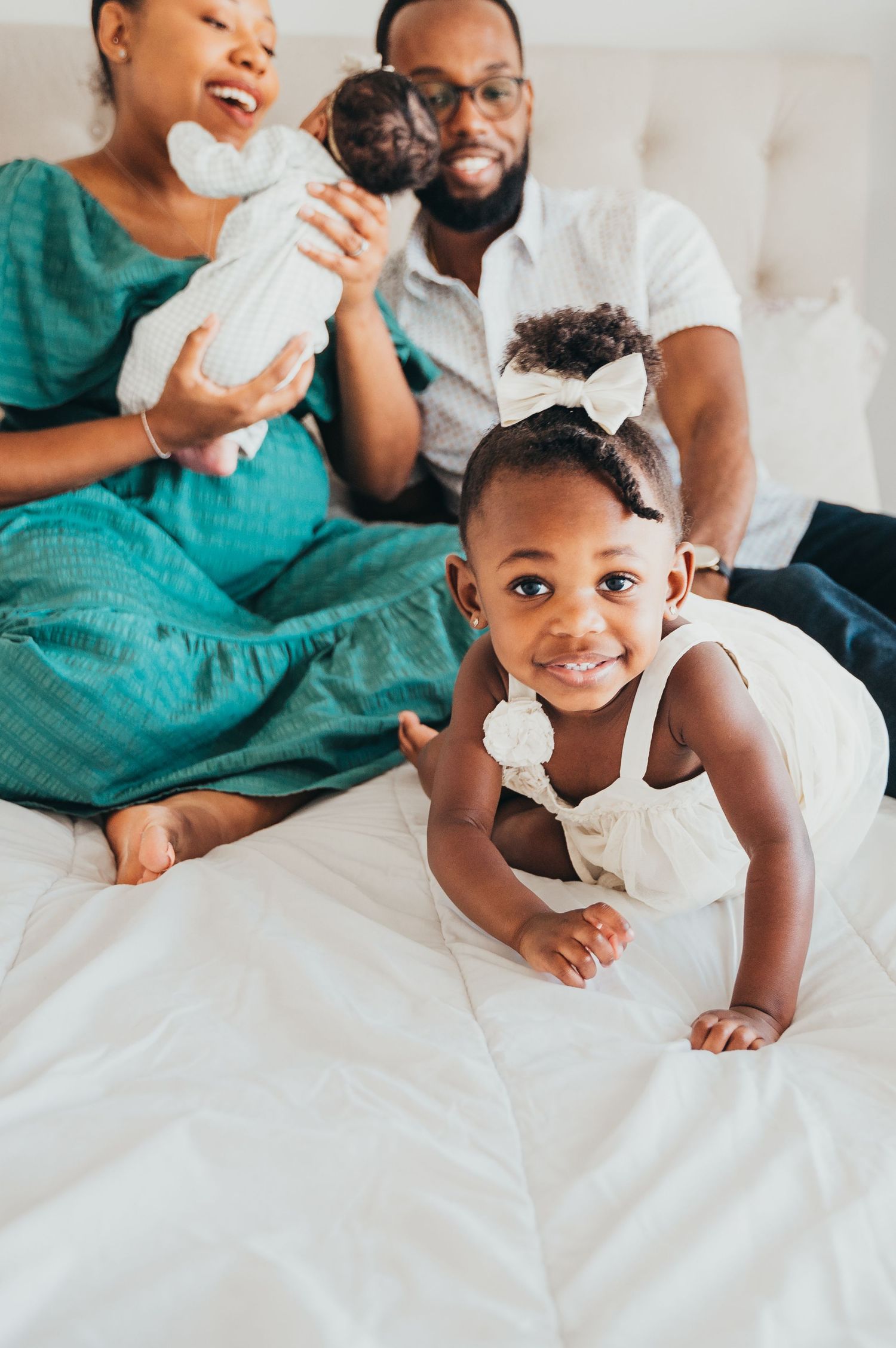 Mom holds newborn and smiles looking at baby and dad looks at toddler crawling toward camera on white bed.