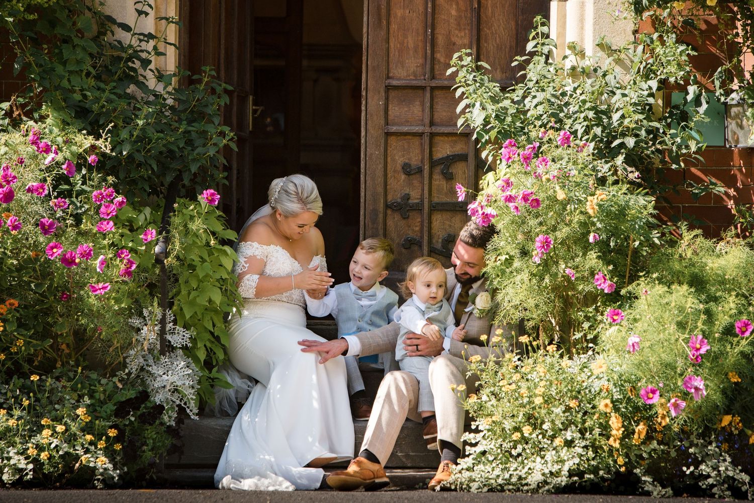 Bride and groom sit on flower-lined steps with two young boys in a sunny outdoor wedding portrait.