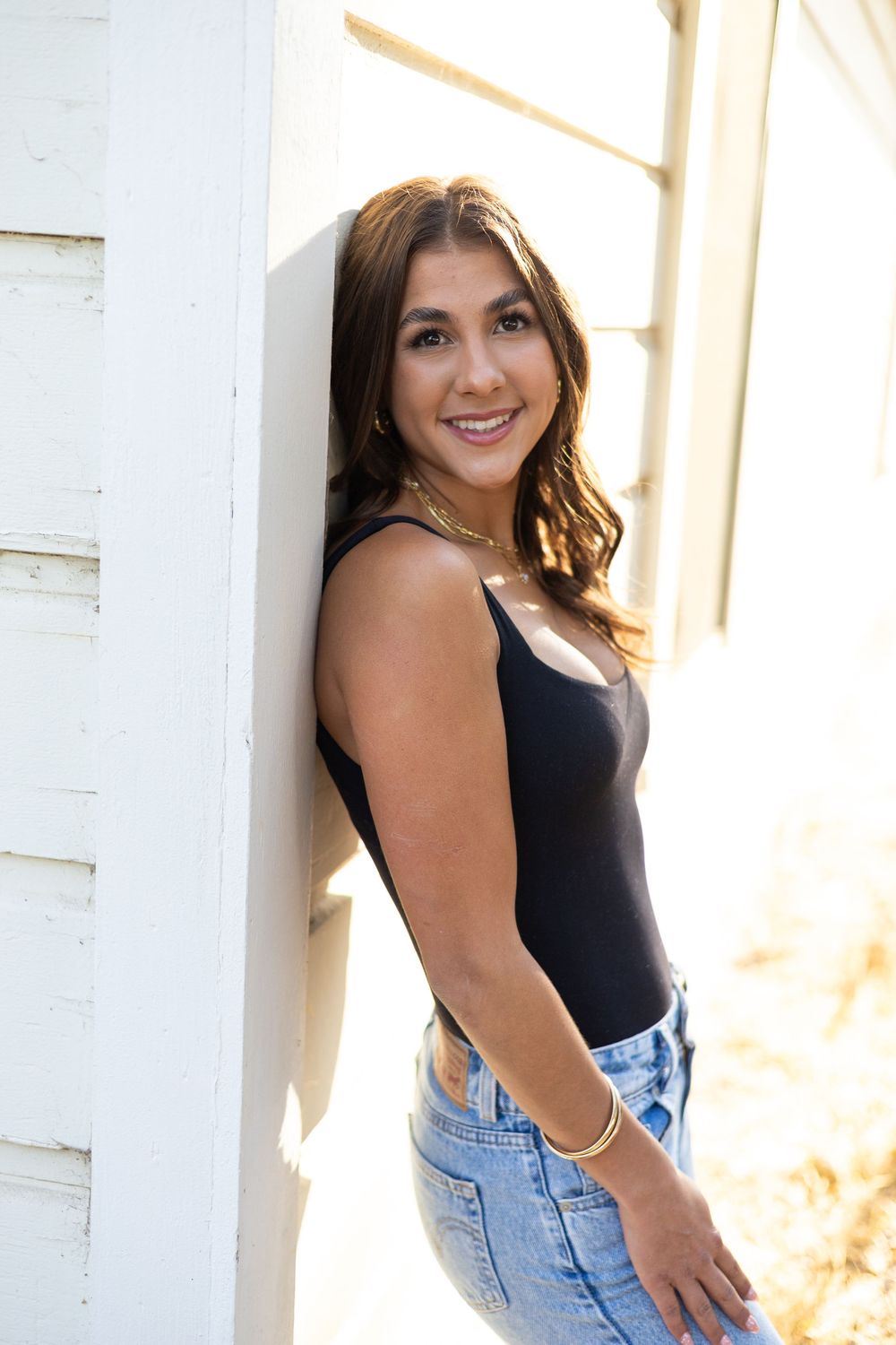 Young woman in black bodysuit and jeans leaning against white wall, smiling confidently in bright natural light.