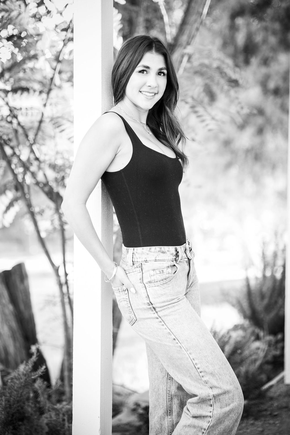 Black and white portrait of a young woman in jeans and tank top leaning against a white pillar outdoors.