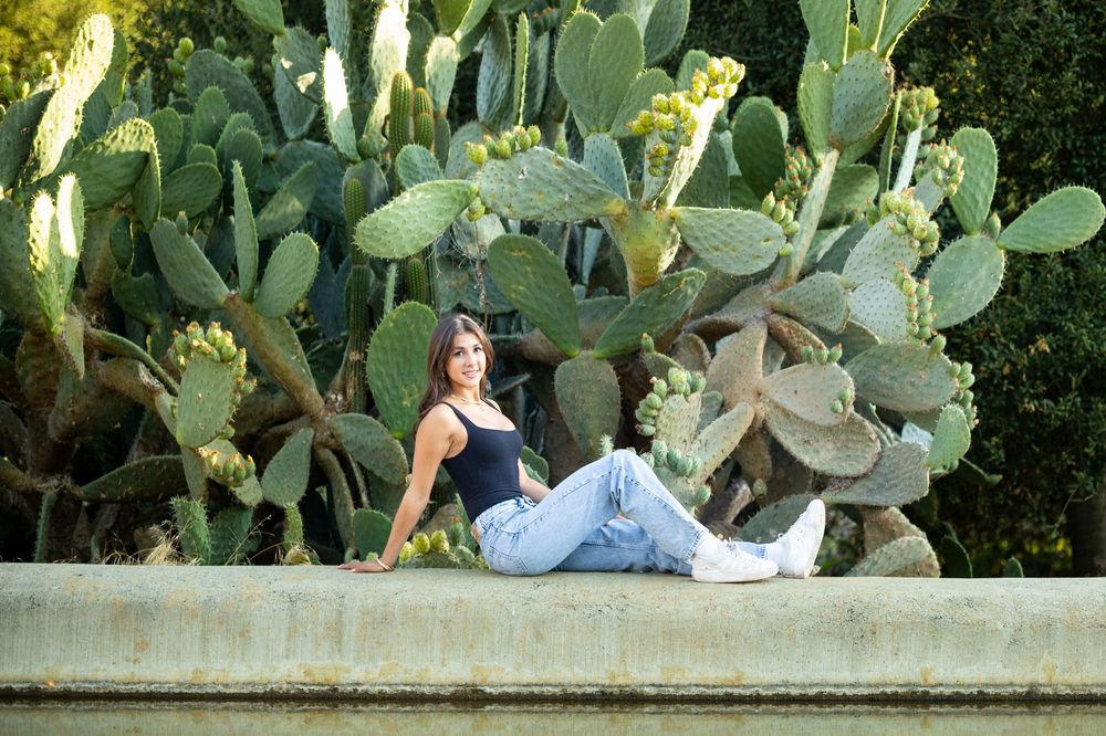 Woman in black top and jeans sitting on a ledge in front of large prickly pear cactus plants outdoors.