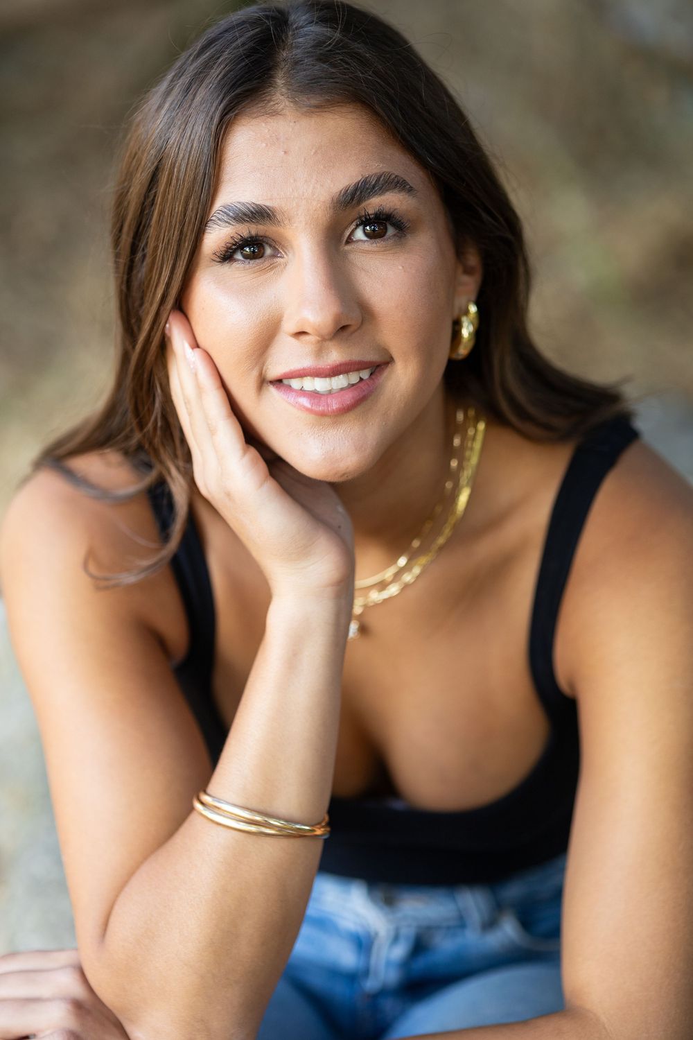 Young woman smiling, wearing gold jewelry and black top, posing with hand on cheek outdoors.