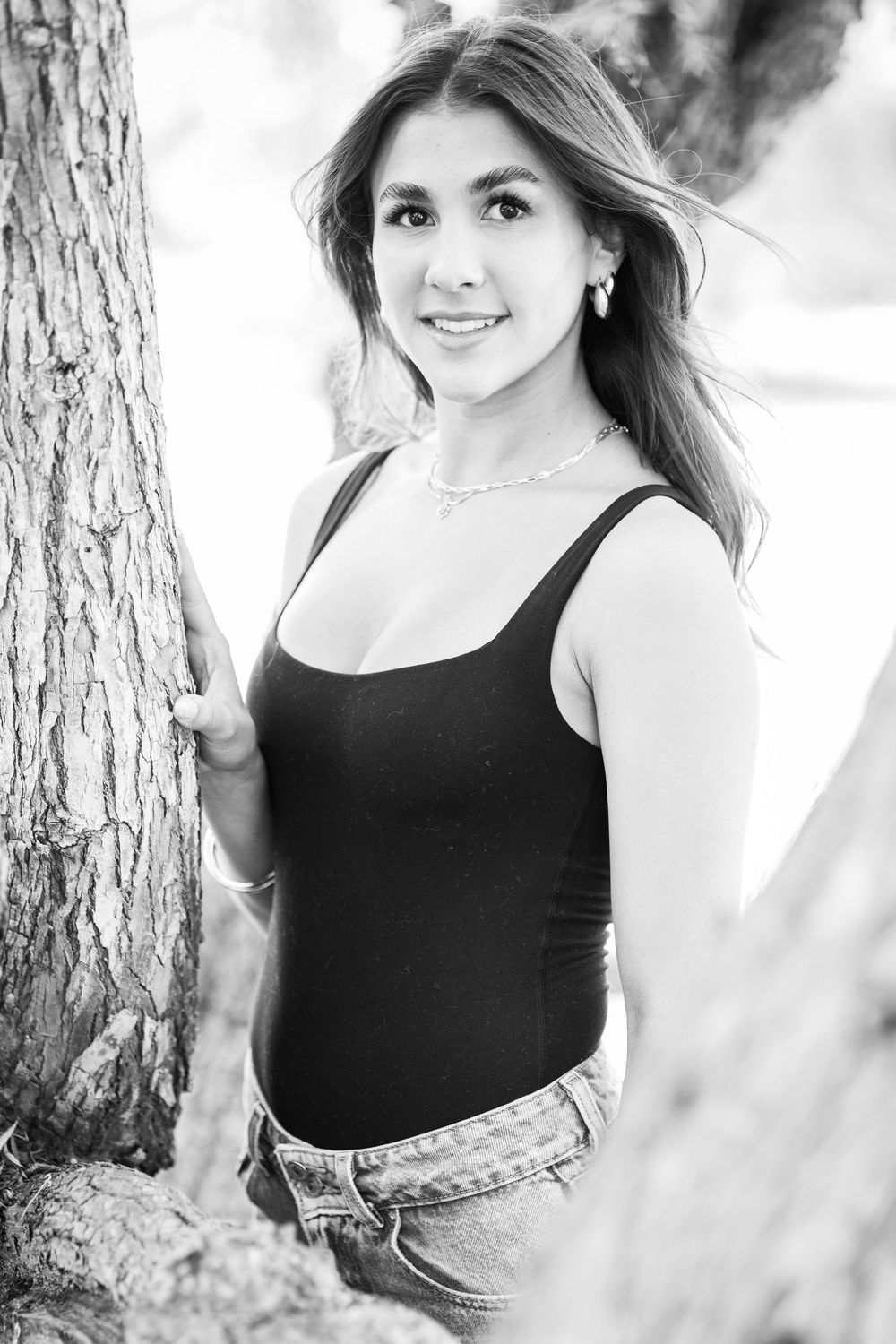 Black and white portrait of a young woman in a tank top and jeans leaning against a tree outdoors.