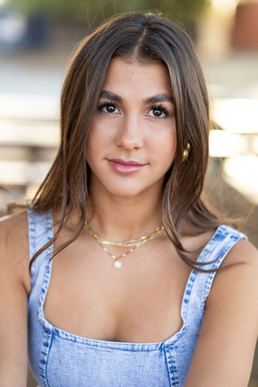 Young woman with brown hair wearing a denim top and gold layered necklaces, posing outdoors in natural light.