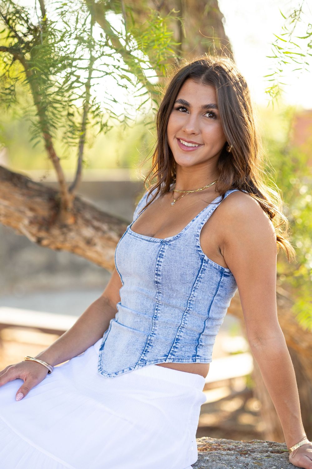 Young woman in denim top and white skirt smiling outdoors among green trees in natural sunlight.