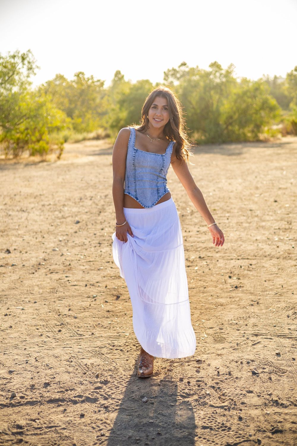 Young woman in blue crop top and white maxi skirt posing in sunny desert landscape with green trees.