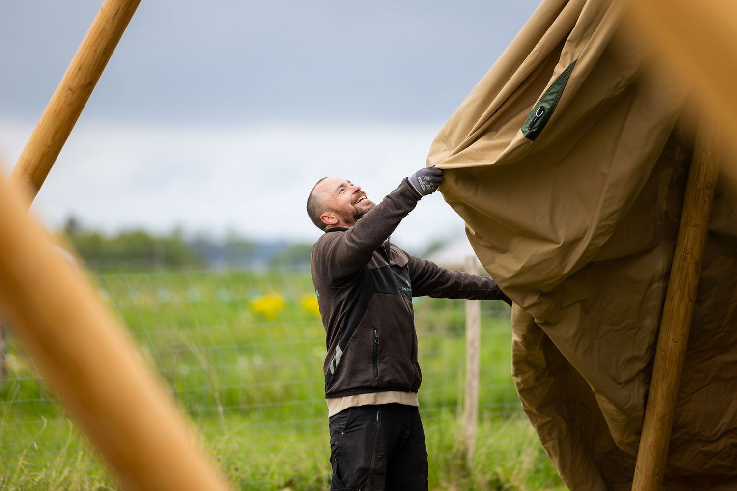 Man setting up a large canvas tent outdoors in a green field on a cloudy day.