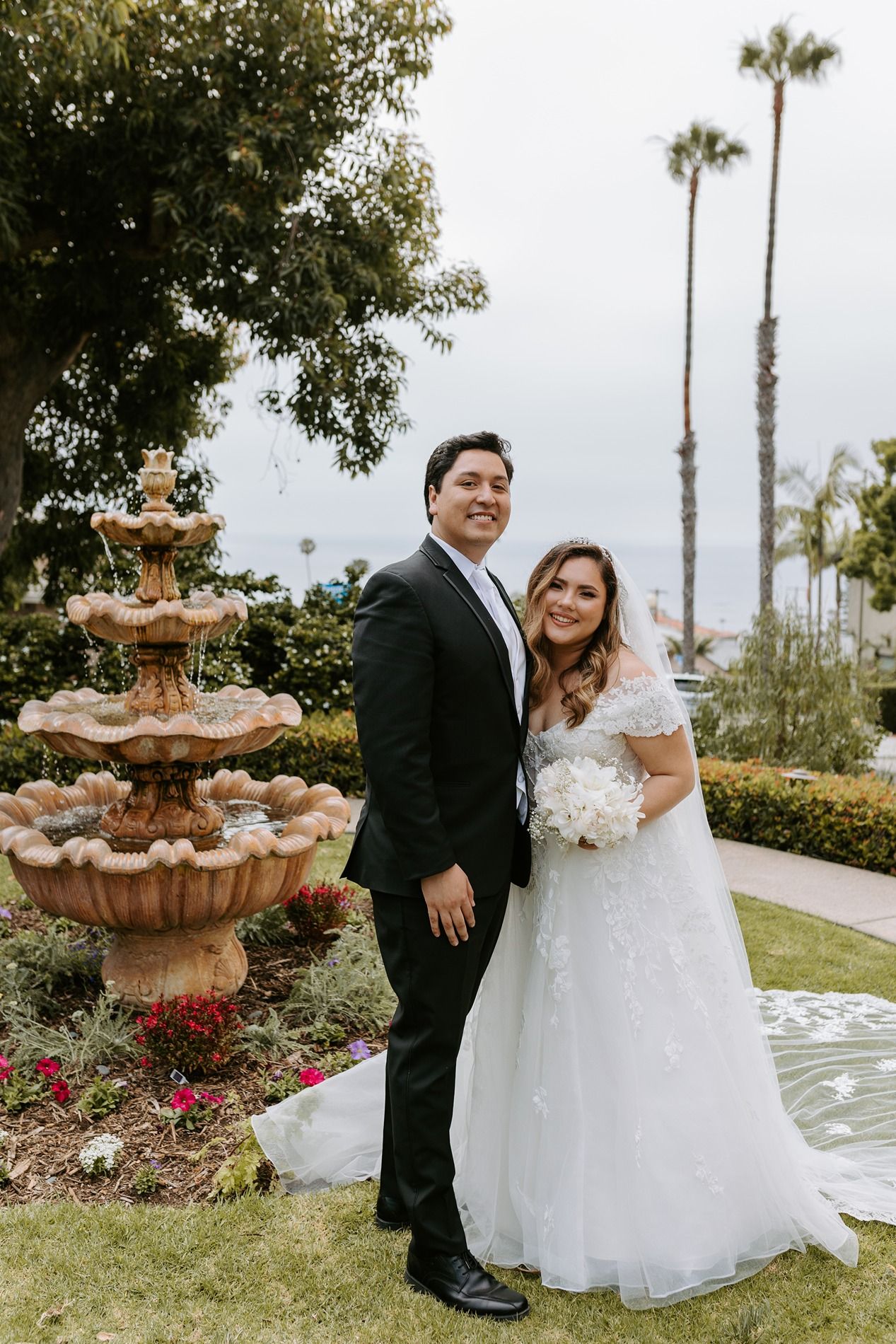 Bride and groom posing together near a stone fountain in a garden, with palm trees and ocean views in the background at The Thursday Club.