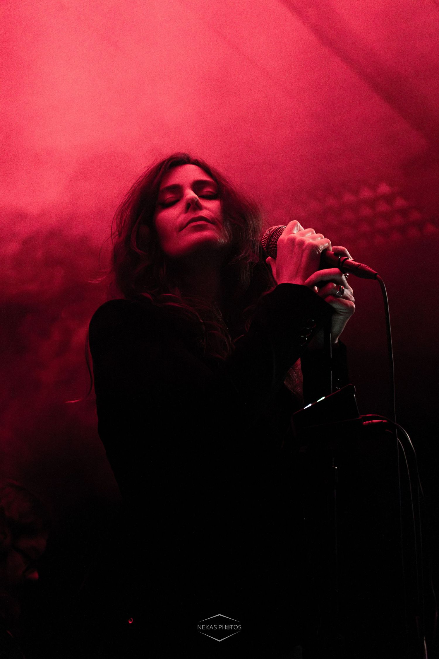 A female singer performs on stage under dramatic red lighting, holding a microphone with eyes closed.