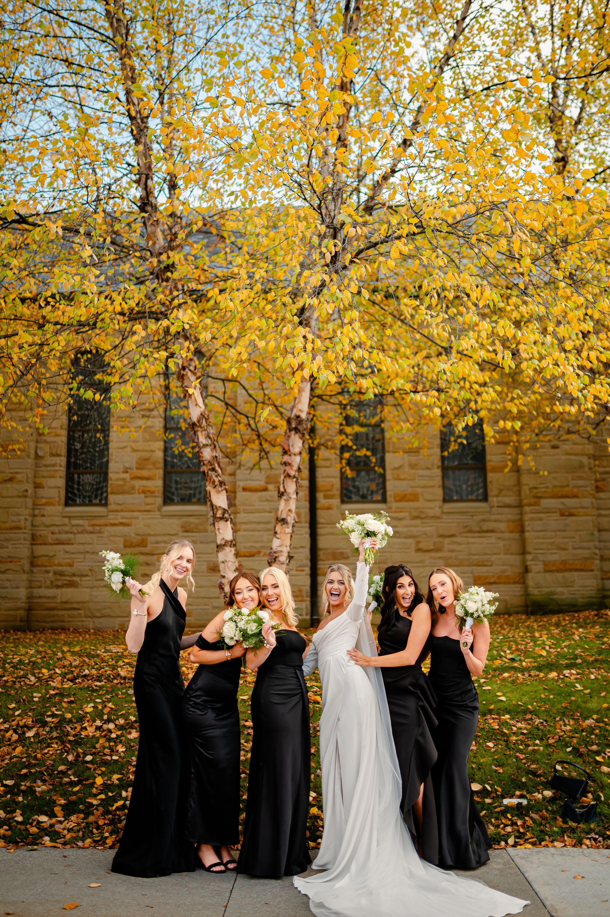 Bride in white gown with bridesmaids in black dresses holding bouquets before autumn tree at stone church wedding.