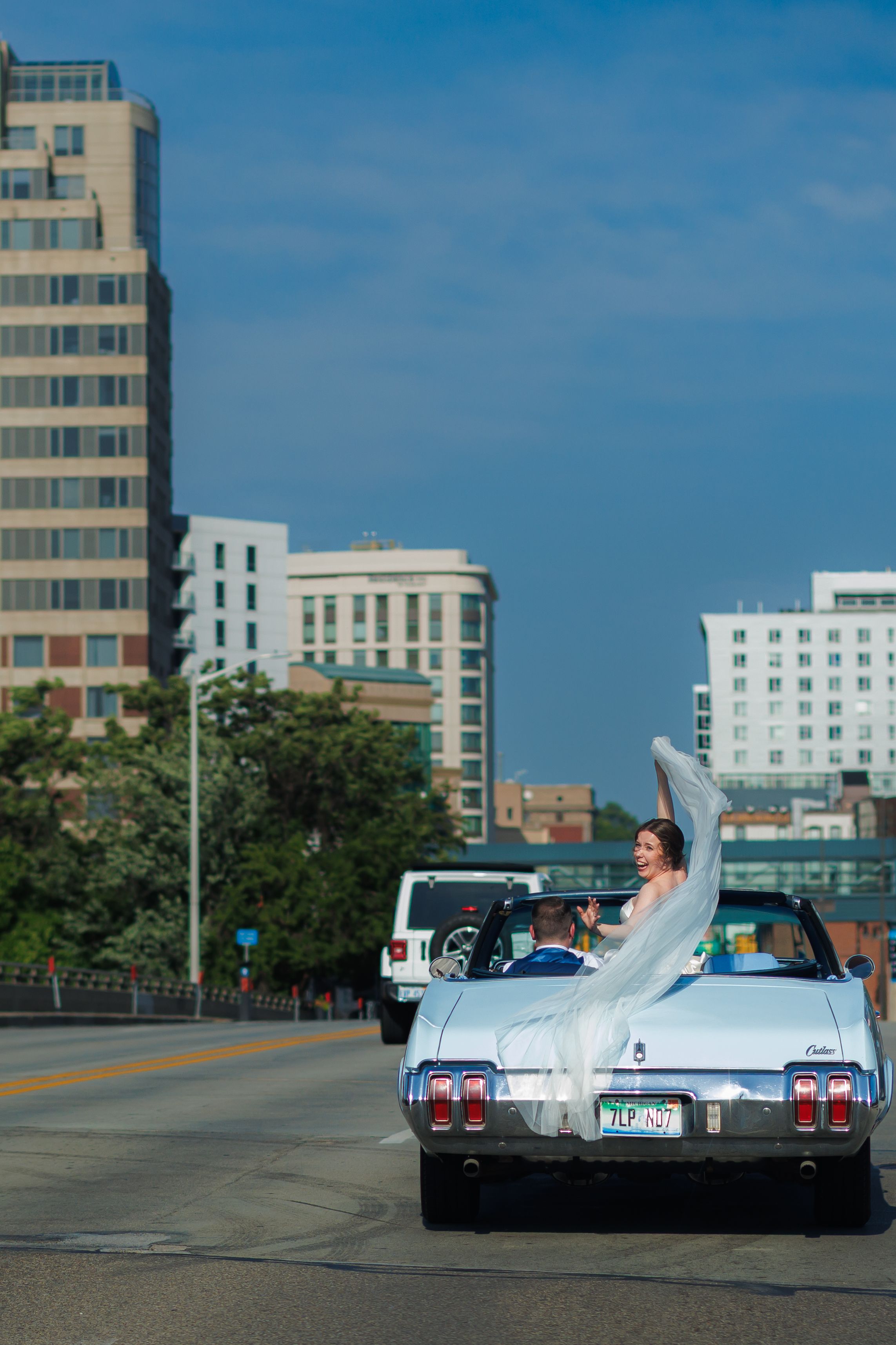 Bride riding in white classic convertible car through city streets on wedding day, veil flowing in wind.