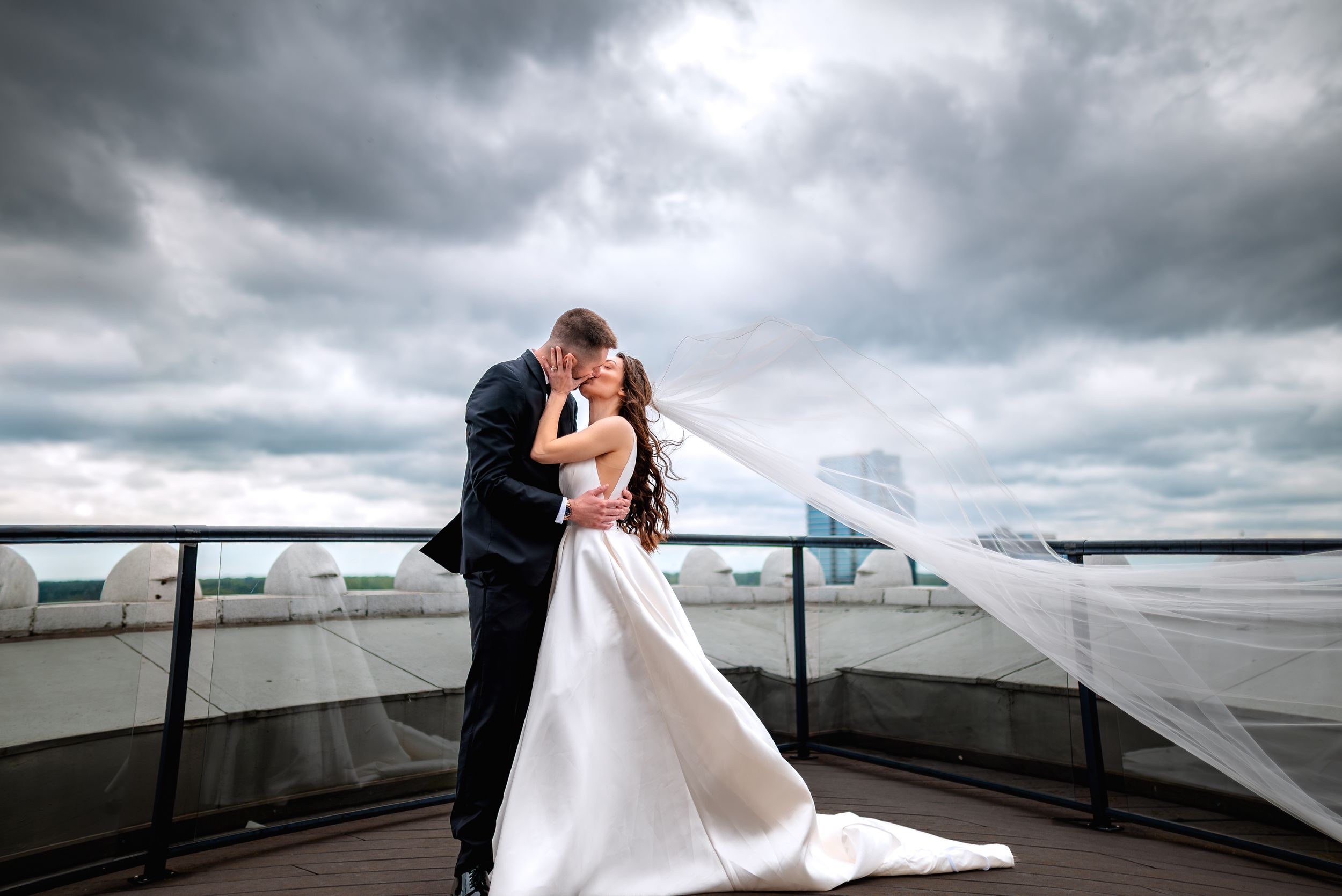 Bride and groom share a romantic kiss on a rooftop, with the wedding veil flowing in the wind under dramatic cloudy skies.