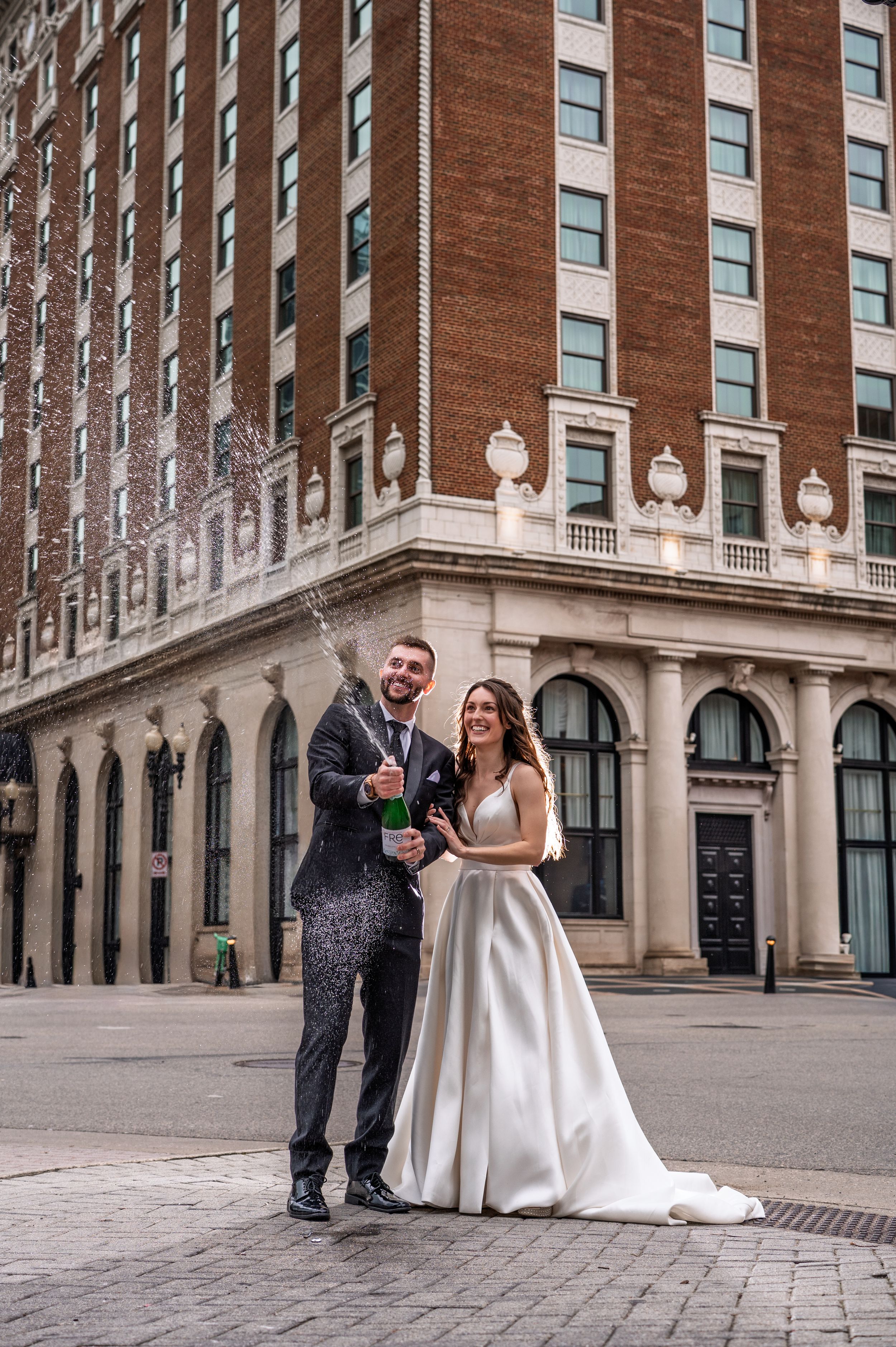 Bride and groom popping champagne outside elegant brick hotel building on their wedding day.