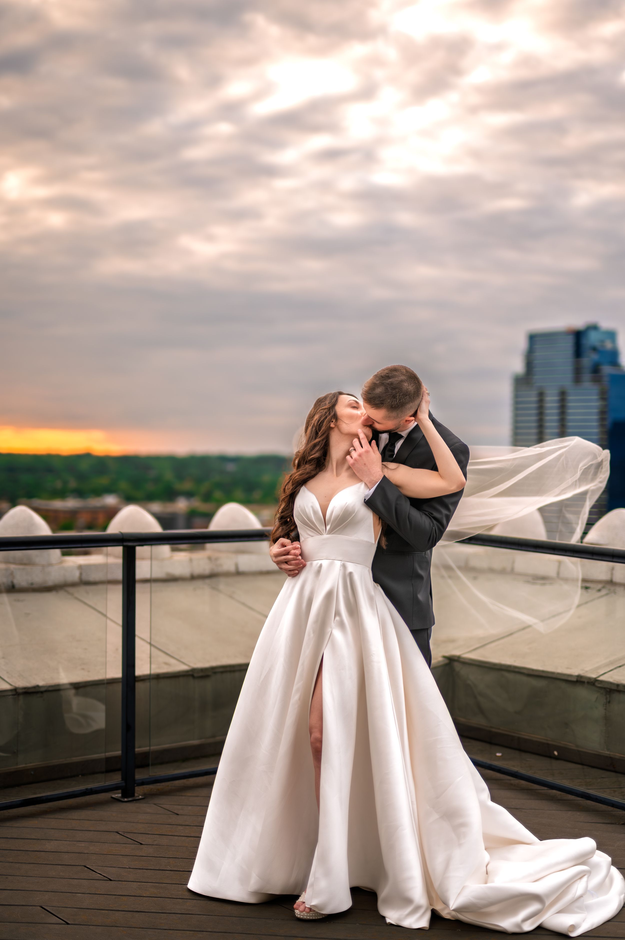 Bride and groom share a romantic kiss on a rooftop at sunset, her white ballgown and veil flowing in the wind.