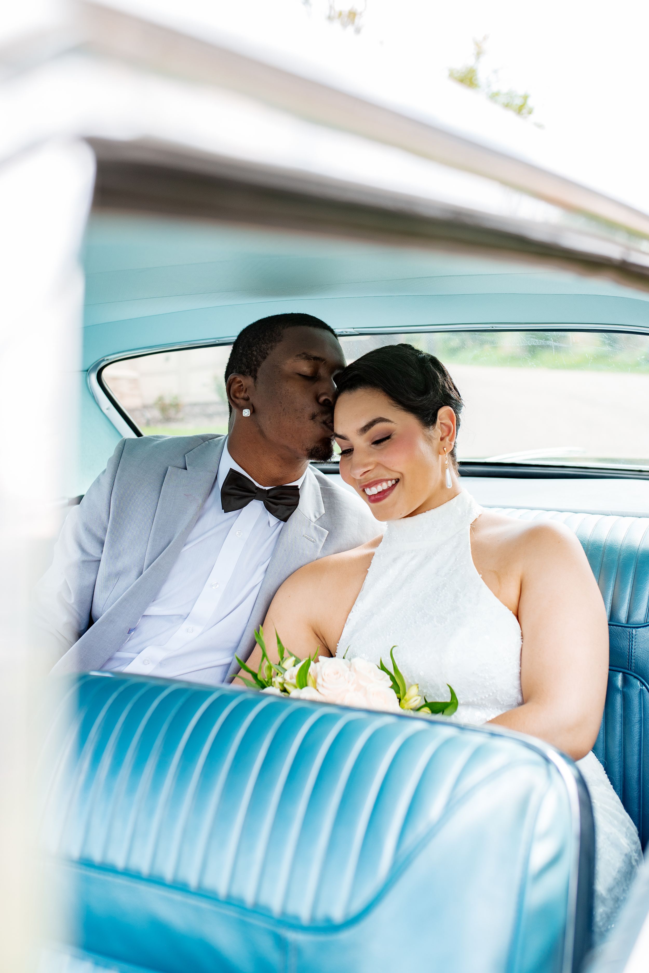 Newlywed couple sharing a romantic moment in a vintage blue car, groom in gray tuxedo kissing bride in white halter gown.