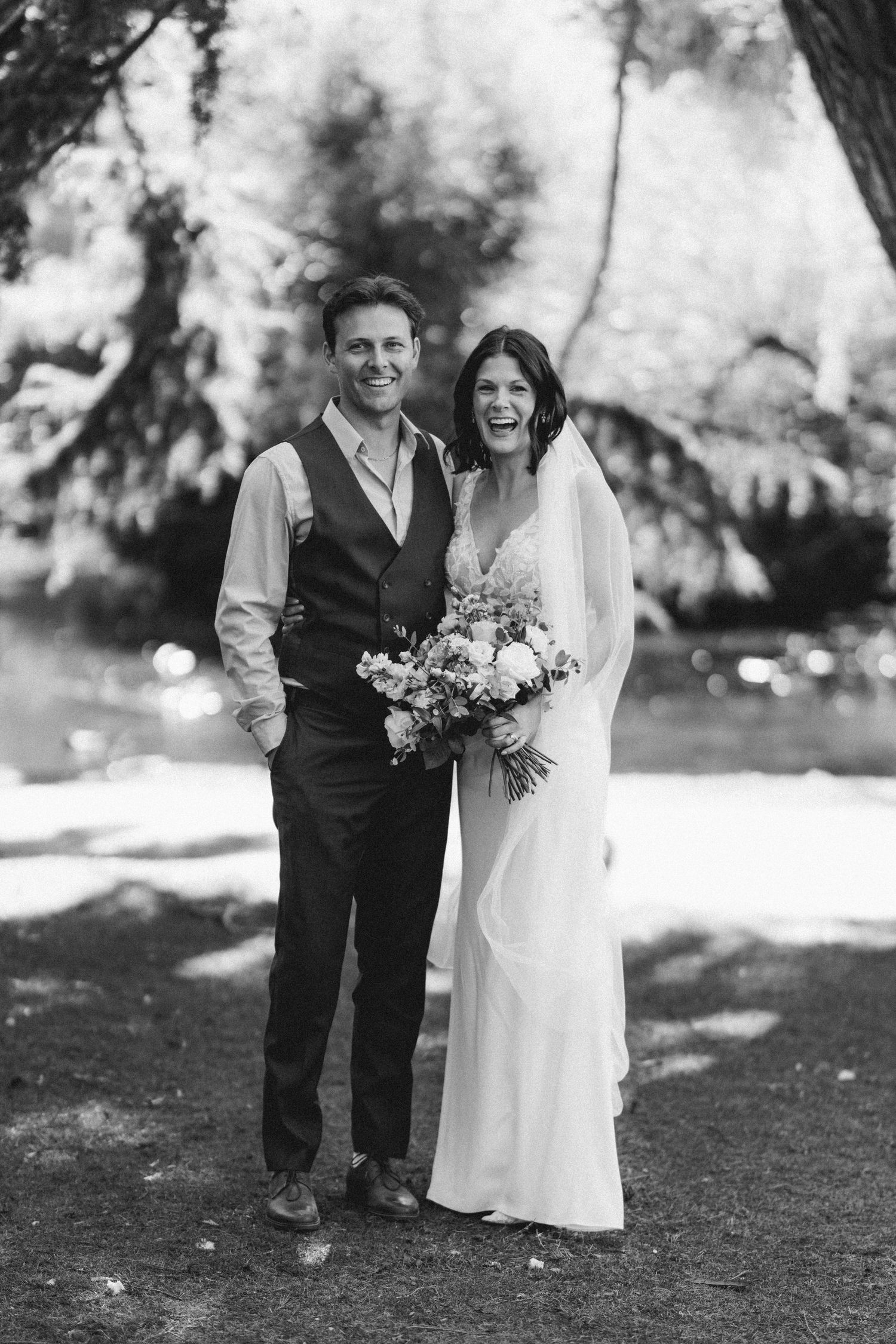 Smiling bride and groom pose together outdoors in black and white wedding photo.