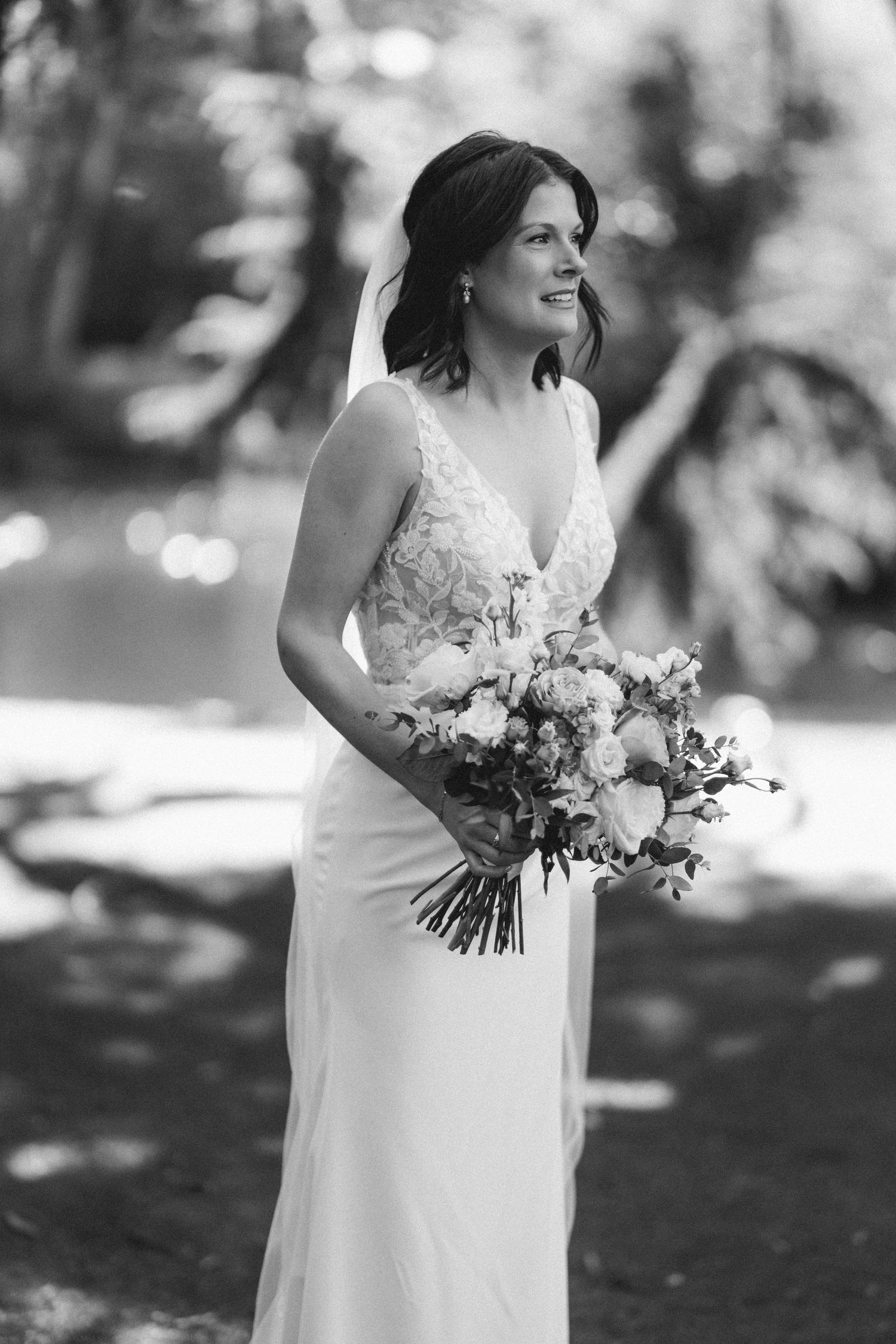 Black and white bridal portrait of bride in lace gown holding bouquet of roses outdoors.