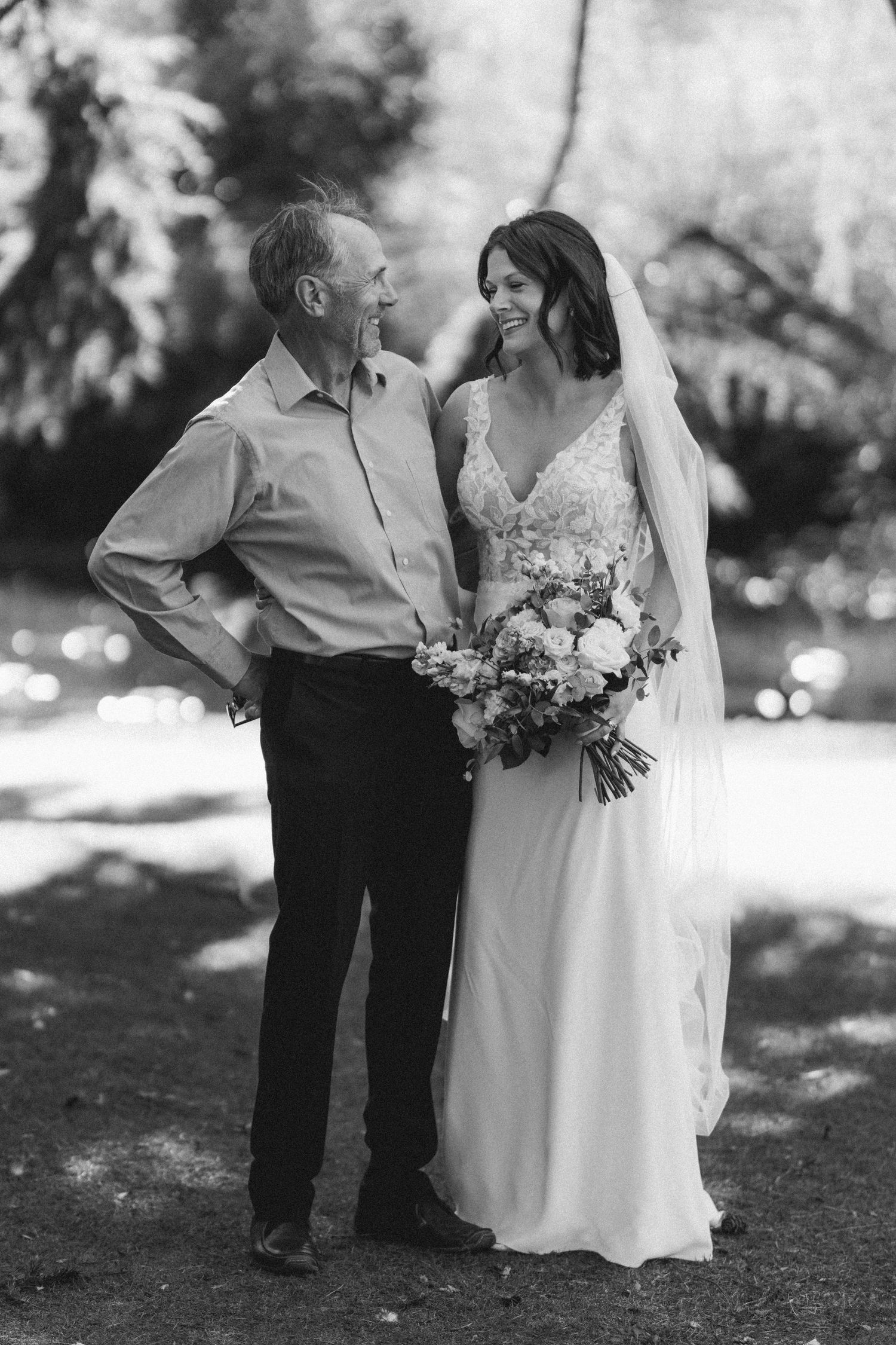Black and white portrait of bride in lace gown with bouquet posing with older man.