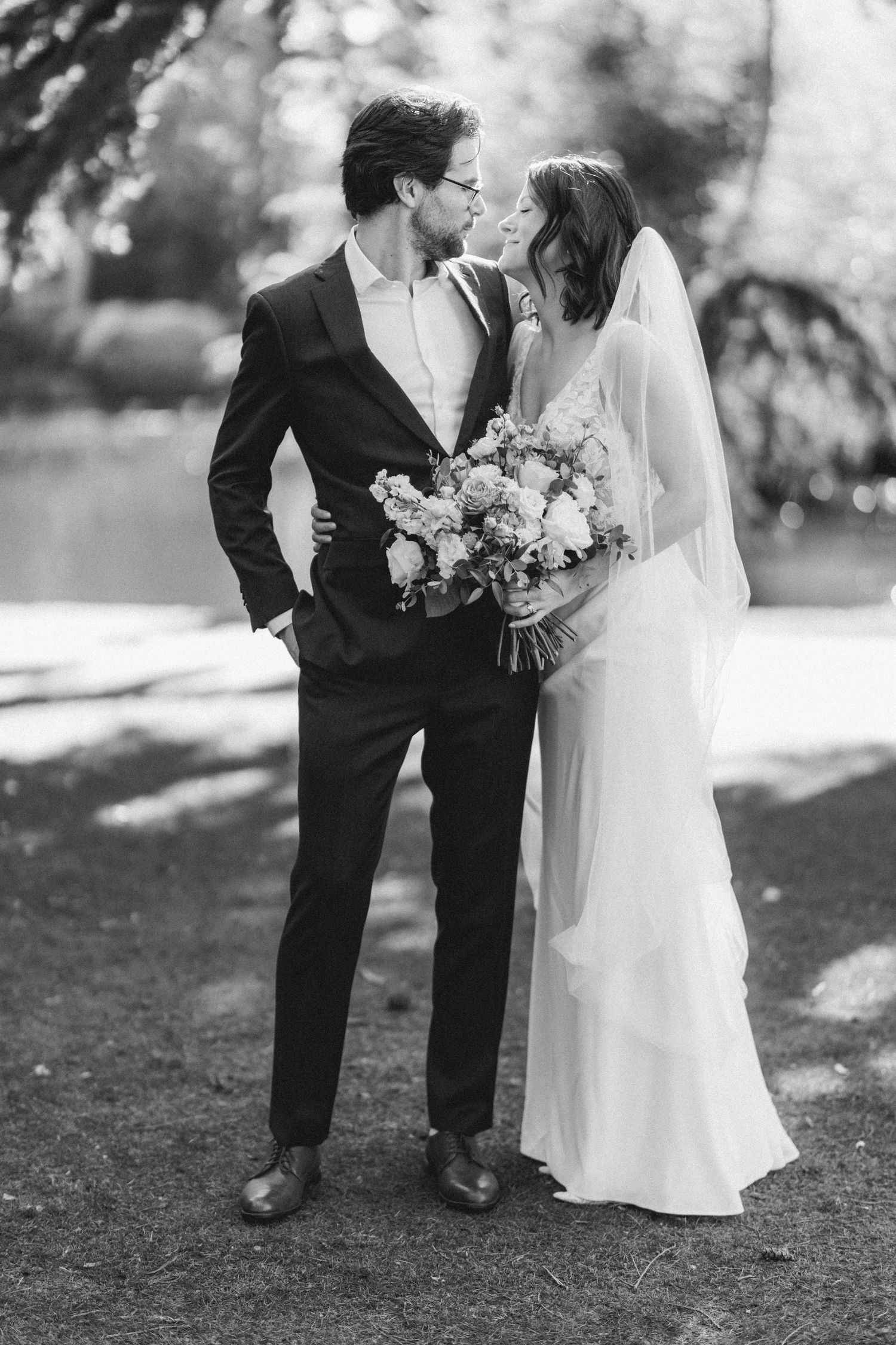Black and white wedding portrait of bride and groom sharing a kiss outdoors near a lake.