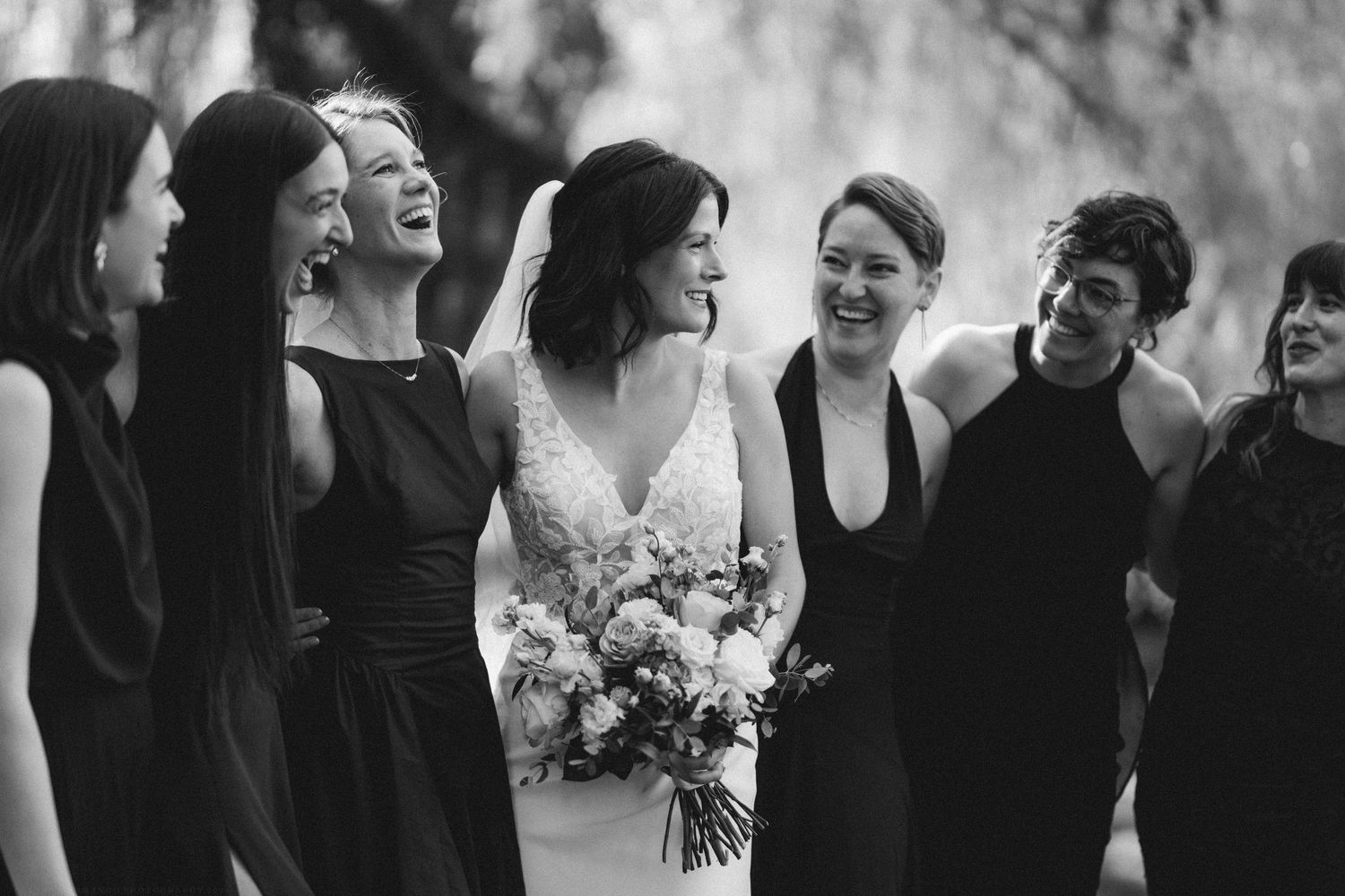 Bride holding bouquet laughs with bridesmaids in black dresses outdoors at wedding.