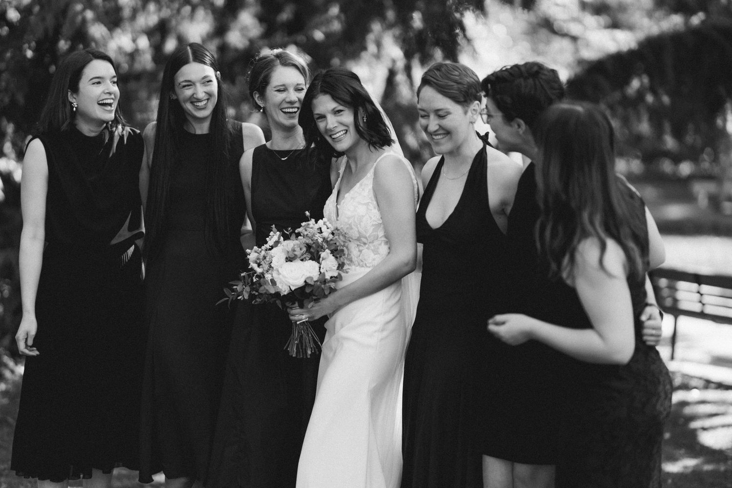 Bride and bridesmaids share joyful laughter together in a black and white outdoor photo.