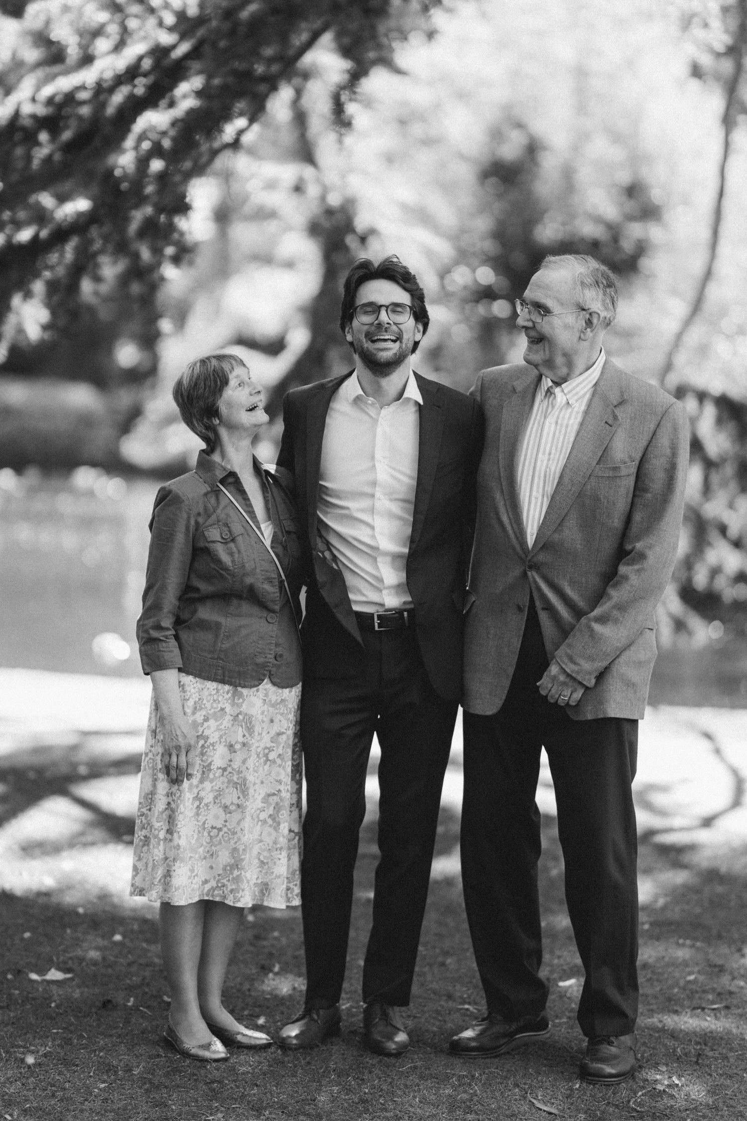 Groom smiles warmly with his parents in a classic black and white outdoor portrait.