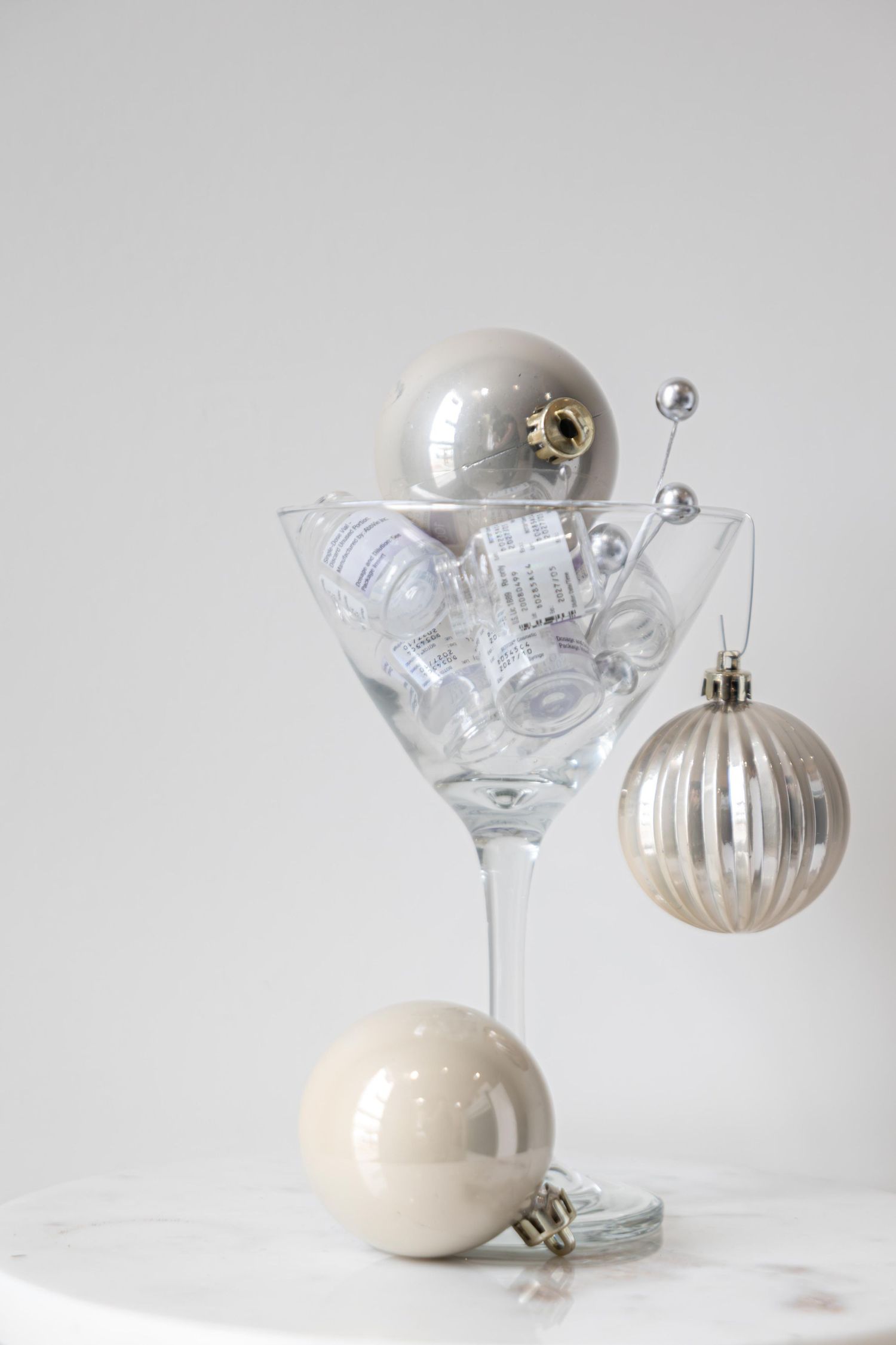 Silver and white Christmas ornaments arranged in a martini glass on a white table for elegant holiday decor.