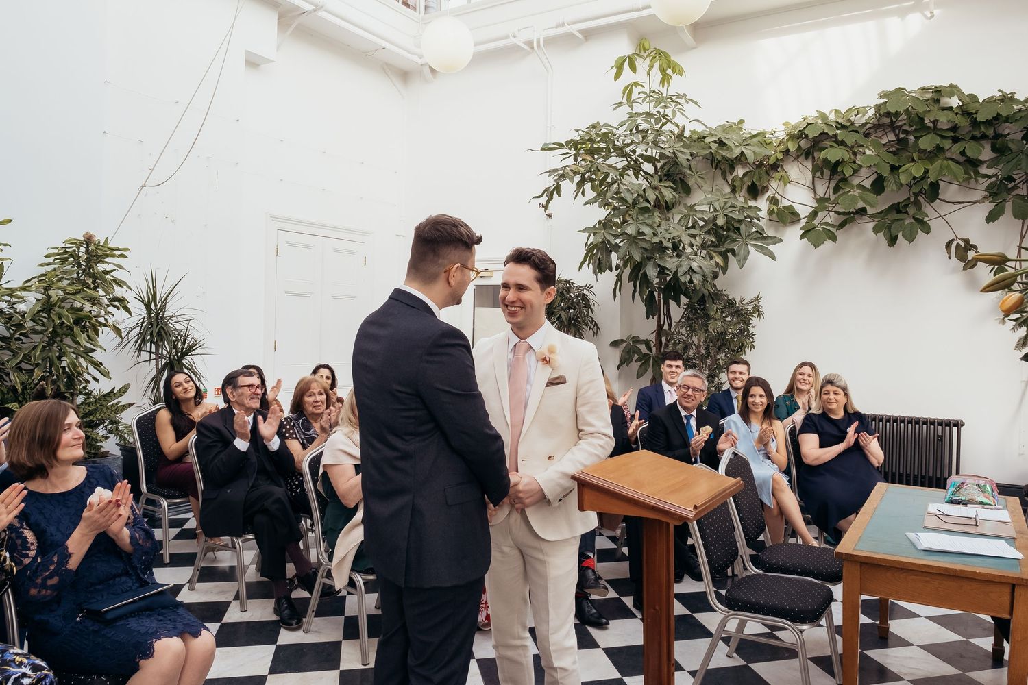 Two grooms exchange vows at a same-sex wedding ceremony on a checkered floor with guests applauding.