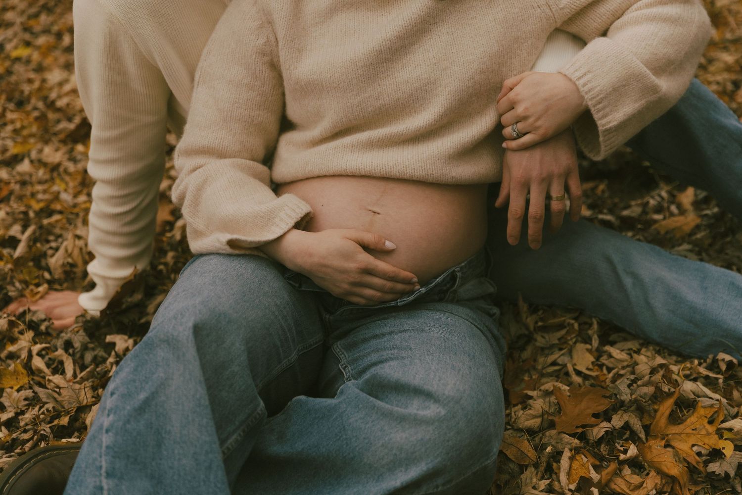 Couple sitting on autumn leaves outdoors, gently cradling a pregnant belly in a warm fall maternity photo.