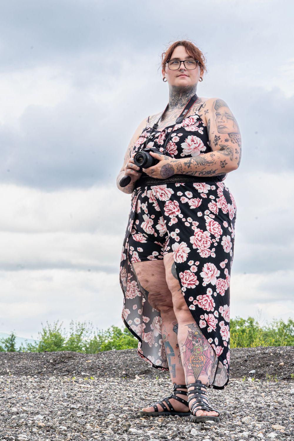 Tattooed plus-size woman in floral dress holding camera outdoors under cloudy sky.