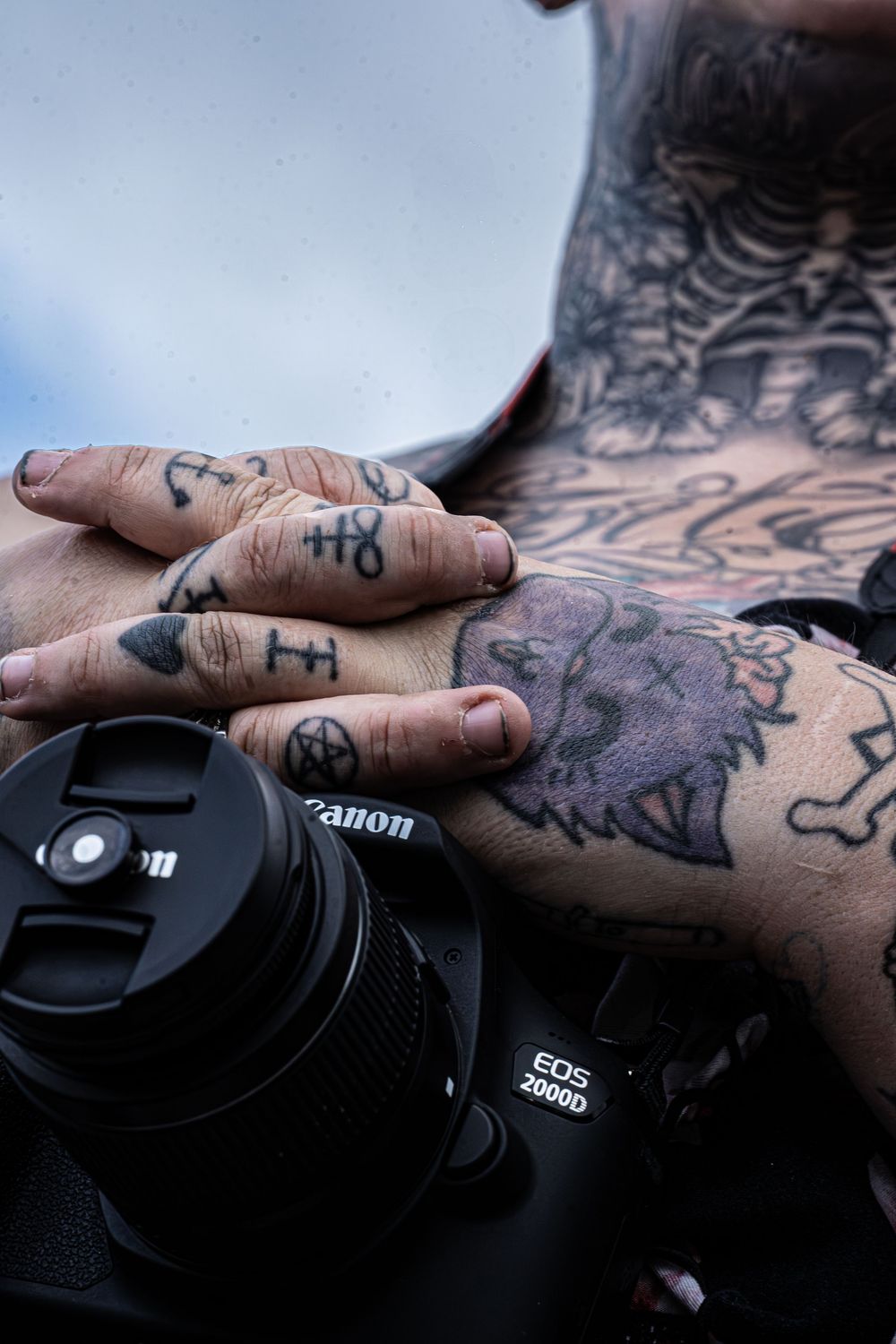 Heavily tattooed hands resting on a Canon EOS 2000D camera, showcasing intricate ink designs on fingers and arms.