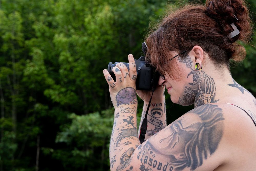 Heavily tattooed woman with gauged ears photographs with a camera outdoors near green trees.