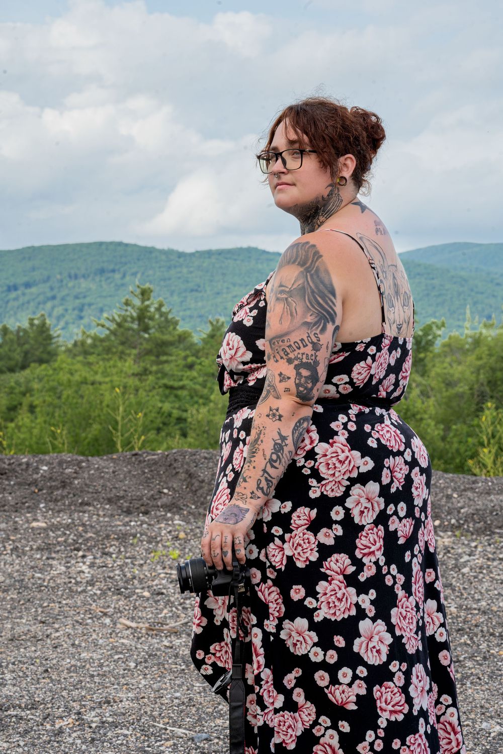 Tattooed woman in floral dress holding camera, posing outdoors with green mountains and cloudy sky in background.