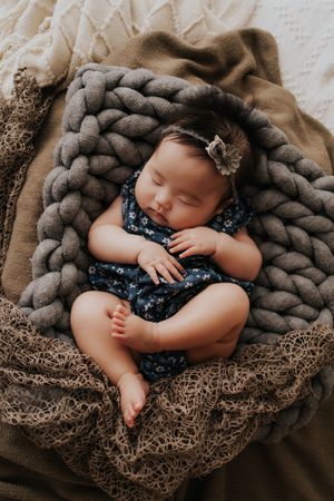 Sleeping newborn baby girl in floral romper and headband nestled in chunky knit blanket.穿著碎花連身衣，戴著髮帶，裹在厚厚的針織毯裡熟睡的新生女嬰。