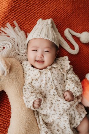 Baby wearing white knit hat and floral onesie smiles while lying on orange knit blanket with plush toy.戴著白色針織帽和碎花連身衣的寶寶躺在橘色針織毯上，懷裡抱著毛絨玩具