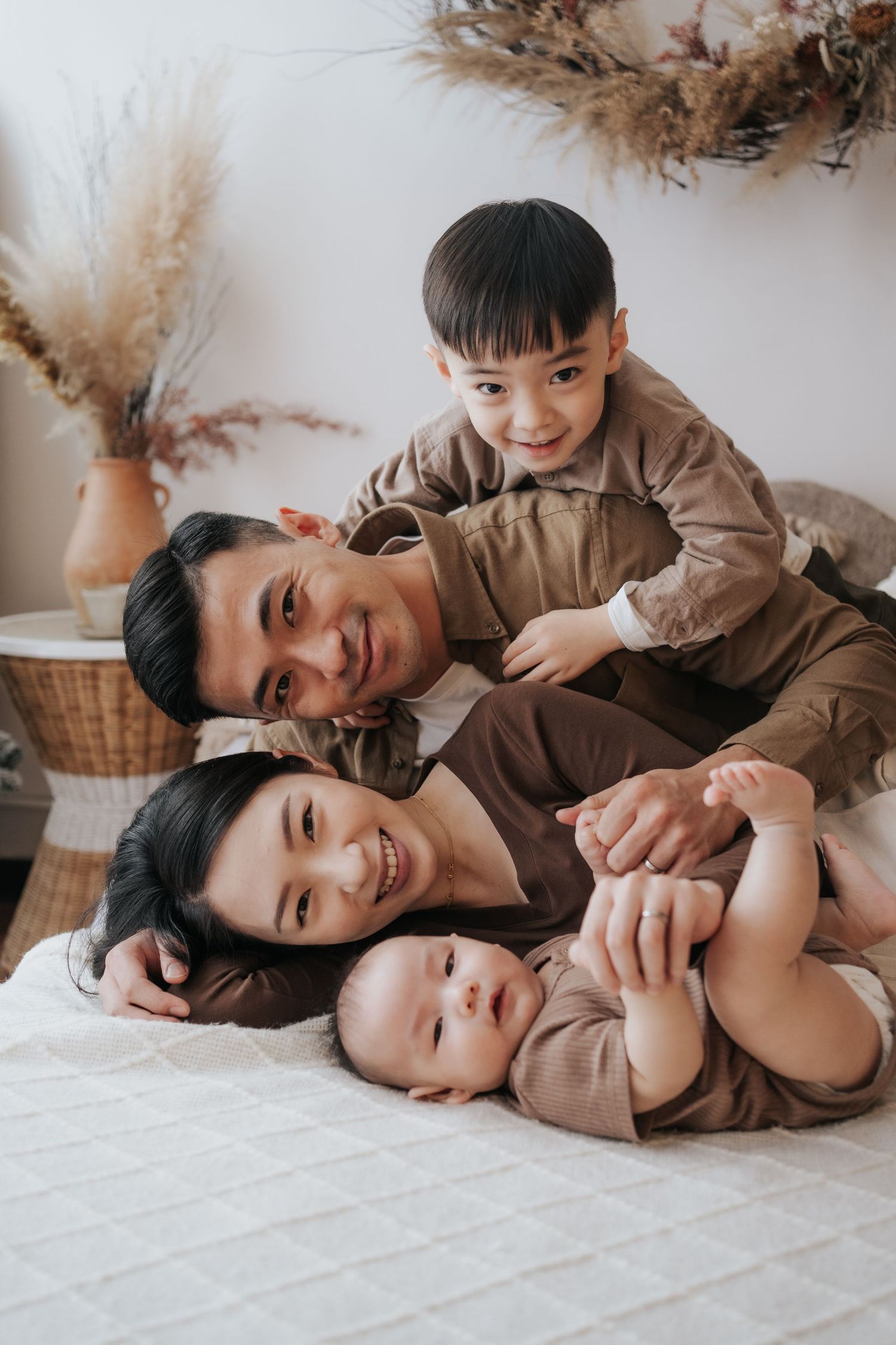 Happy Asian family of four posing together on a bed, parents smiling with toddler and baby in matching brown outfits.幸福一家四口在床上合影，父母面帶微笑