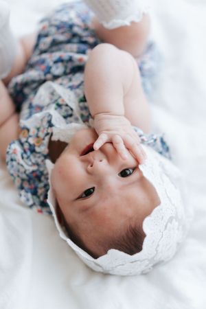 A baby wearing a floral dress lies on white bedding while looking up at the camera.一個穿著碎花洋裝的嬰兒躺在白色床單上，抬頭看著鏡頭。