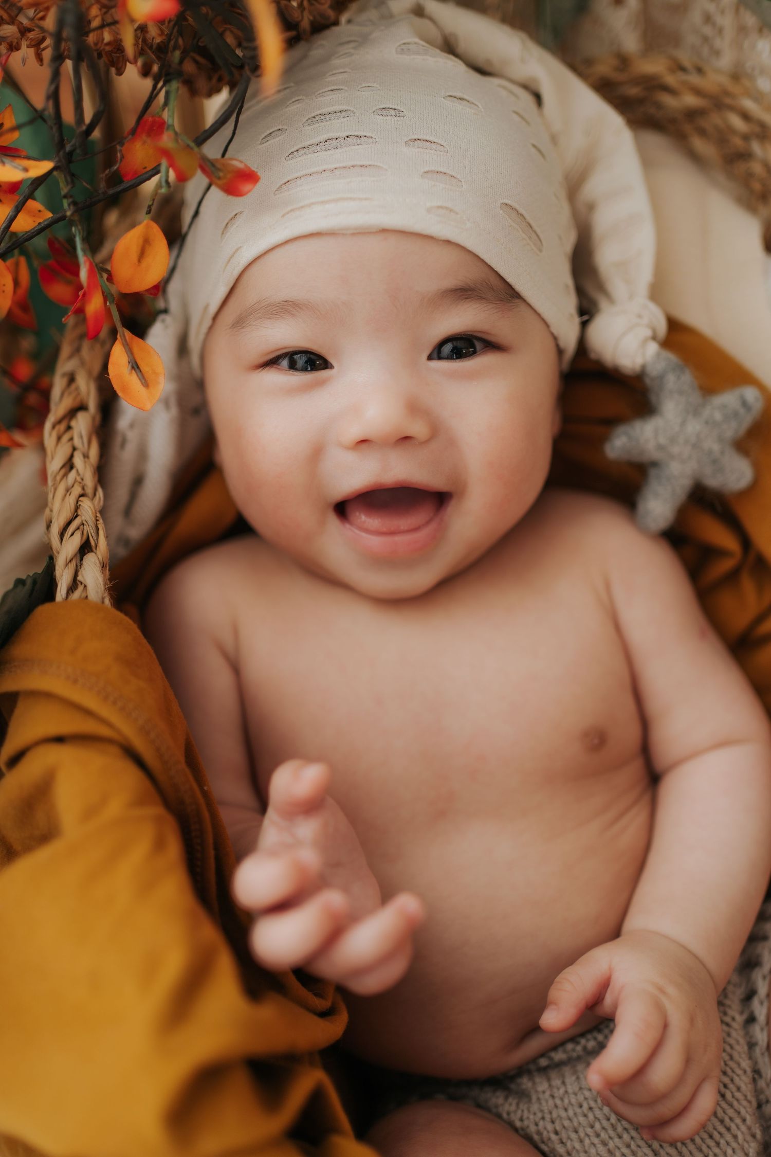 A joyful baby wearing a cream beanie lies on soft textiles surrounded by autumn-colored decor and orange flowers.一個戴著奶油色毛線帽的快樂嬰兒躺在柔軟的紡織品上