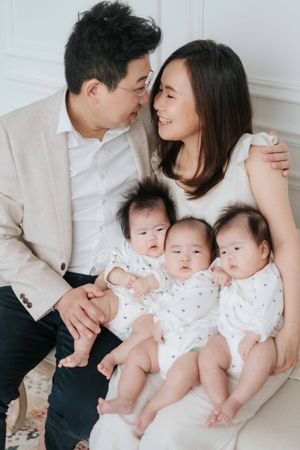 Asian couple smiling at each other while holding three adorable babies in white onesies during a family portrait session.夫婦抱著三個穿著白色連身衣的可愛寶寶
