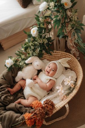 Baby in white knit outfit lying in woven basket with floral decorations and plush toy surrounded by orange flowers.身穿白色針織服的嬰兒躺在編織籃中，籃子上裝飾著鮮花