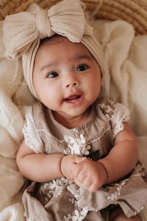 Sweet baby wearing a beige bow headband and lace dress smiles while lying on cream colored bedding.可愛的寶寶戴著米色蝴蝶結髮帶，穿著蕾絲連身裙，躺在奶油色的床單上微笑。